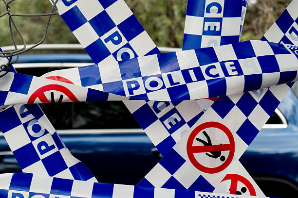 Cordon tape of the New South Wales Police Force covering a hole which has been cut in a fence at Sydney Kingsford Smith Airport.  In the background is one of the cars in the carpark. This image was taken on Ross Smith Avenue, Mascot on a sunny and partly cloudy afternoon on 1 November 2025.
