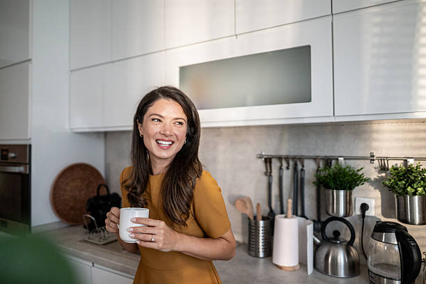 Young woman in a bright modern kitchen smiling while holding a warm mug, enjoying a peaceful morning routine that suggests relaxation, healthy lifestyle and cozy home comfort