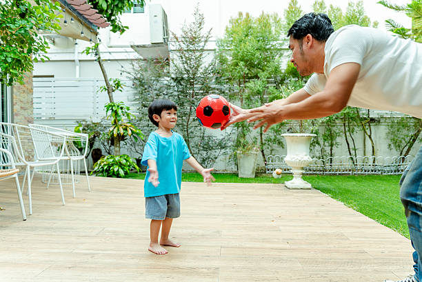 istock Father playing catch with young Asian boy using a red soccer ball in a backyard garden setting. 2244571788