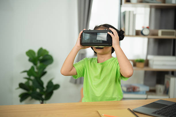 istock Child Engaged in Virtual Reality Experience with VR Headset while Sitting at Table in Modern Room with Laptop and Indoor Plant in Background 2244572874