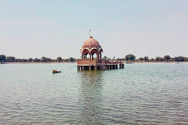Jaisalmer, Rajasthan, India - 22 July, 2024: A weathered stone pavilion emerges gracefully from the calm waters of Gadisar Lake in Jaisalmer, Rajasthan, India. Featuring a domed roof, ornate arches, and sculpted pillars, this historic monument reflects ancient Indian architecture and cultural heritage. Birds perch along the pavilion’s railing while a small boat rests nearby, creating a serene and photogenic scene ideal for travel, tourism, and landscape photography. The tranquil waterfront and sunlit reflections highlight the timeless charm of this architectural landmark.