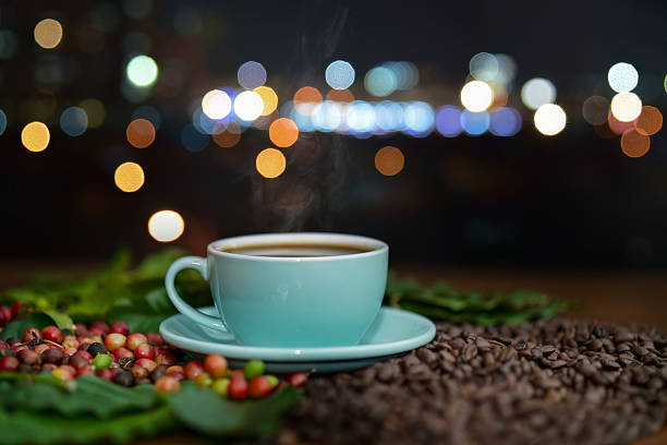 Steaming cup of freshly brewed black coffee in light blue cup sits surface covered with roasted coffee beans surrounded by coffee cherries and vibrant green leaves.