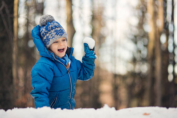 Young boy wearing a blue winter jacket and knit hat with pom-pom laughs joyfully while holding a freshly packed snowball in his raised hand. His face radiates pure delight with an open-mouthed smile and sparkling eyes as he prepares to throw the snowball. Bare winter trees creating are blurred in the background. Golden afternoon light filters through the forest.Shot with Canon R5