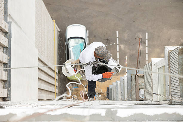 An ironworker on top of a steel building frame. The young Hispanic man is climbing on a roof beam wearing a hardhat, attached to a safety harness.