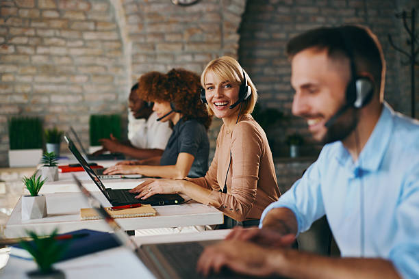 istock Smiling Caucasian female customer service agent looks directly at the camera while working on her laptop with diverse colleagues in a busy, modern call center office 2244588970