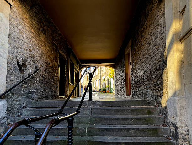 A narrow, stone-walled alleyway in Frome, Somerset, known as Sheppards Barton. The view is from a dark, arched passage looking out into a sunlit, cobblestone street. The buildings lining the street are made of stone and feature windows with white frames. Potted plants are visible on the street, and two people can be seen walking in the distance.