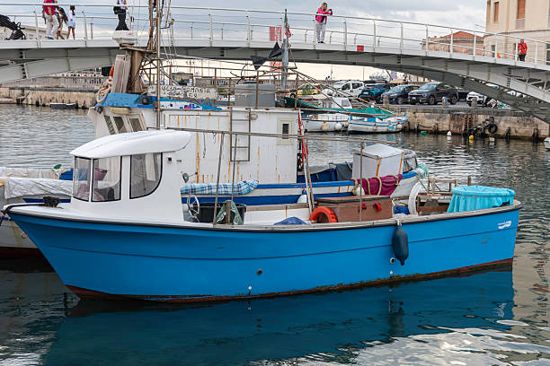 Syracuse, Sicily, Italy - October 20, 2025: Fishing boats moor at a charming harbor in Syracuse, Sicily, Italy, calm waters, and urban surroundings. Capturing a vibrant coastal Italian scene with traditional boats and docks.