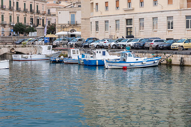 Syracuse, Sicily, Italy - October 20, 2025: Fishing boats moor at a charming harbor in Syracuse, Sicily, Italy, calm waters, and urban surroundings. Capturing a vibrant coastal Italian scene with traditional boats and docks.