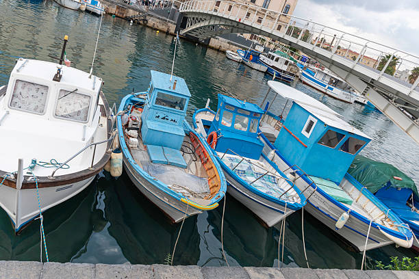 Syracuse, Sicily, Italy - October 20, 2025: Fishing boats moor at a charming harbor in Syracuse, Sicily, Italy, calm waters, and urban surroundings. Capturing a vibrant coastal Italian scene with traditional boats and docks.