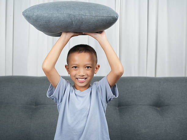 Cheerful boy holds gray pillow above his head, ready for playful moment. background features soft, neutral colored couch and curtains, creating cozy atmosphere