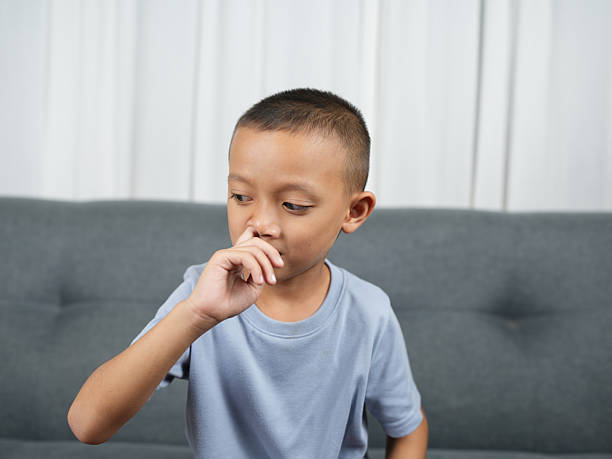 istock Young boy with short hair is sitting on gray couch, looking thoughtful while touching his nose. His casual blue shirt complements neutral background, creating serene atmosphere 2244607788