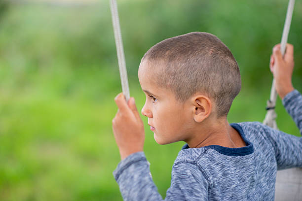 A young boy with a shaved head sits on a swing in a lush green park, gazing into the distance. A calm, reflective outdoor moment capturing childhood and play.