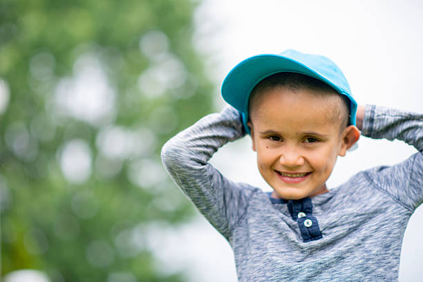 A cheerful young boy in a blue cap and gray long-sleeve shirt stands outdoors, smiling as he adjusts his hat. Bright, sunny day with blurred greenery in the background.