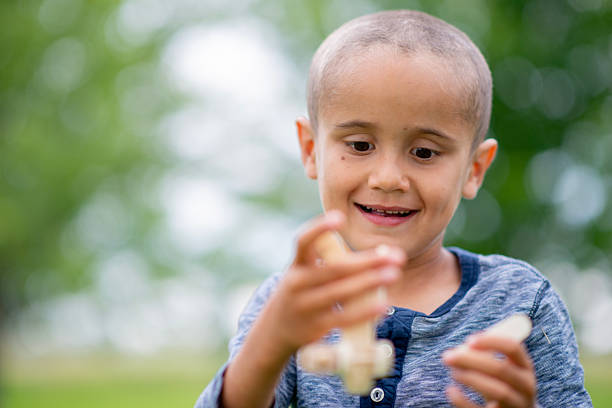 A cheerful Asian boy wearing a blue shirt enjoys a sunny day at the park, playing with a wooden toy airplane. Lush greenery and soft bokeh background emphasize curiosity and playful discovery.