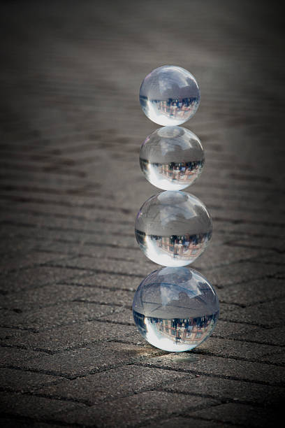 Four clear crystal photography balls balanced vertically on dark, paved ground, inverting and reflecting the surrounding urban landscape and blue sky.