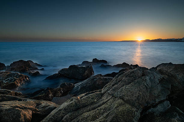 Long exposure of twilight on the coast of Gran Canaria with volcanic rocks and calm ocean water. Warm colors on the horizon blend with deep blue sky.