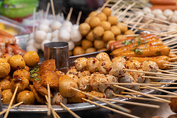 Assorted street food skewers and fried snacks on a circular grilling tray at a market.