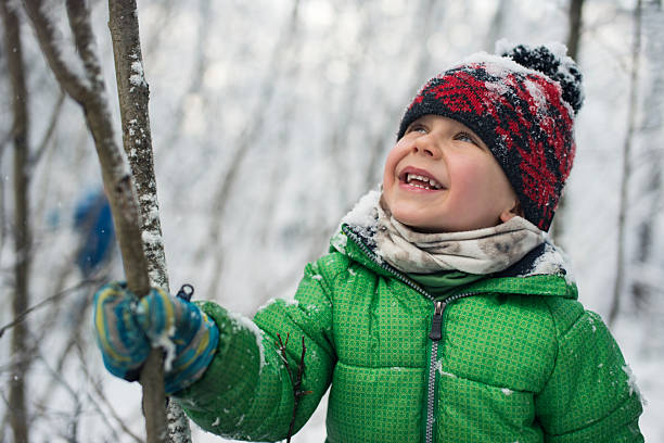 Young boy wearing a bright green winter jacket grips a thin tree trunk and tilts his head back in laughter as he shakes it to release snow from the branches above. His face radiates pure delight, mouth open in genuine laughter as the disturbed snow falls around him, with flakes visible on his hat and clothing.Shot with Nikon D800