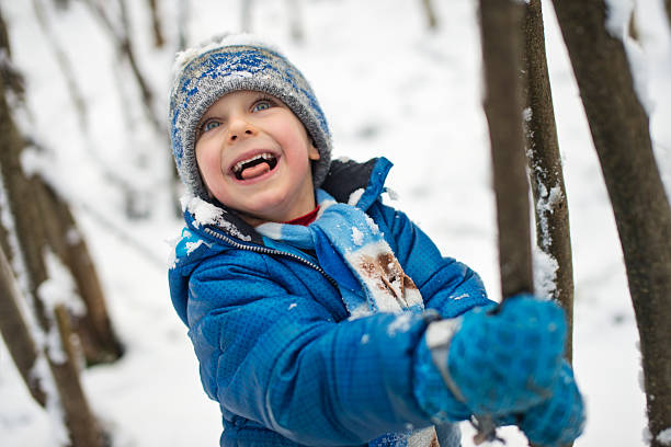 Young boy wearing a blue winter jacket grips a thin tree trunk and tilts his head back in laughter as he shakes it to release snow from the branches above. His face radiates pure delight, mouth open in genuine laughter as the disturbed snow falls around him, with flakes visible on his hat and clothing.Shot with Nikon D800
