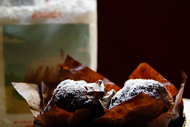 American Double Extra Chocolate Muffin Sprinkled with Powdered Sugar Still Life, Silhouette of Bag of Powdered Sugar in the Background (Left), Famous Hungarian “Koronás Porcukor” (in English, Crown Powdered Sugar) of “Koronás Cukor” Brand (in English, Crown Sugar Brand) by Hungary’s Largest Sugar Refinery (Since 1894) in Kaposvar, Somogy County of Austrian Agrana Group (in Vienna, Since 1988), Indoors, Only Sunlight from Window (Budapest, Hungary, Central and Eastern Europe - September 9, 2025)
