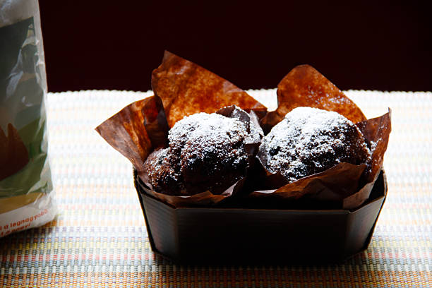American Double Extra Chocolate Muffin Sprinkled with Powdered Sugar Still Life, Silhouette of Bag of Powdered Sugar in the Background (Left), Famous Hungarian “Koronás Porcukor” (in English, Crown Powdered Sugar) of “Koronás Cukor” Brand (in English, Crown Sugar Brand) by Hungary’s Largest Sugar Refinery (Since 1894) in Kaposvar, Somogy County of Austrian Agrana Group (in Vienna, Since 1988), Indoors, Only Sunlight from Window (Budapest, Hungary, Central and Eastern Europe - September 9, 2025)