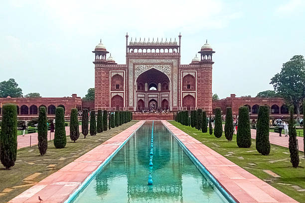 Agra, Uttar Pradesh, India - 25 July, 2024: A monumental red sandstone Mughal gateway ends a long reflecting pool, flanked by trimmed trees and manicured lawns. Visitors explore a heritage site framed by timeless architectural beauty in India.