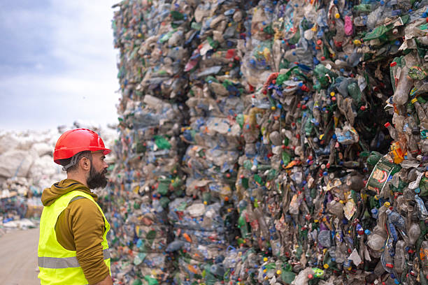 istock Man Wearing Red Helmet Inspecting Large Stacked Plastic Bottle Bales At Recycling Centre 2244643220