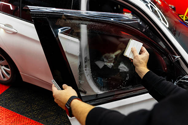 istock Detailer apply dark tint film to car side windows using a squeegee for smooth, bubble-free adhesion. Smoothing dark tint film on side windows removes air bubbles and ensures perfect alignment. 2244646545