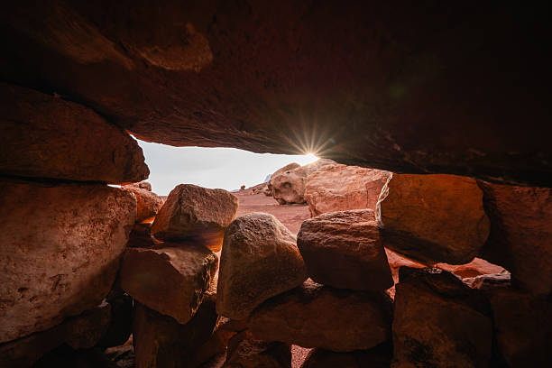 The setting sun shines through a gap in stacked red rocks in a desert landscape.
