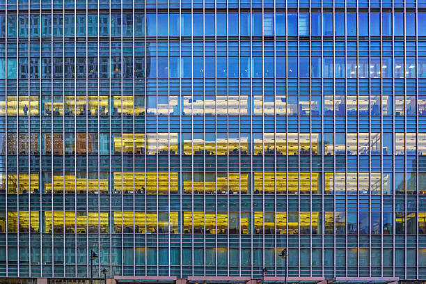 A full-frame, eye-level shot captures the repetitive, grid-like facade of a modern skyscraper, defined by blue glass windows and metal frames, where several horizontal floors glow with bright golden-yellow interior office lighting, contrasting with the unlit blue panes above and below. This detailed architectural pattern is perfect for conveying themes of finance, corporate life, business, technology, late work hours, or abstract urban environment design.