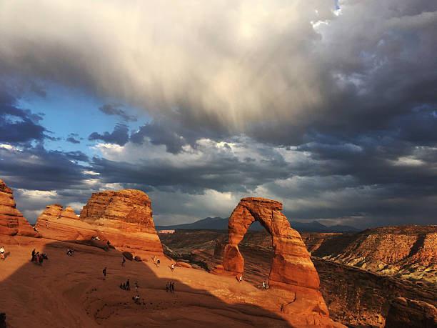 Moab, UT, USA - 5 July 2018: People have hiked to Delicate Arch in Arches National Park as the sun sets at the Golden Hour. Sweeping dramatic clouds and golden light wash over this famous arch.