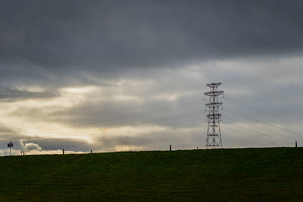 Electric power transmission tower standing on green hill under dramatic cloudy sky illustrating energy distribution infrastructure, renewable connection, and modern electricity network concept