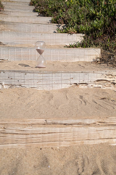 Hourglass placed on sandy wooden steps with green foliage in the background, representing the concept of passing time, transitions, and the flow of life's journey