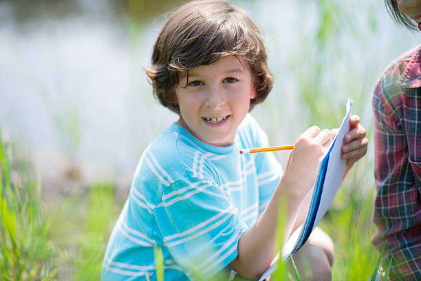 A cheerful boy sits in a sunlit field, writing and drawing on a clipboard with a pencil. This outdoor learning scene conveys curiosity, education, and nature-inspired study.