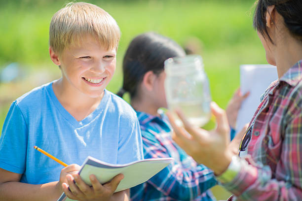 A cheerful boy writes in a notebook while an adult supervisor demonstrates with a jar, as other children observe during an outdoor science activity in a sunny setting.