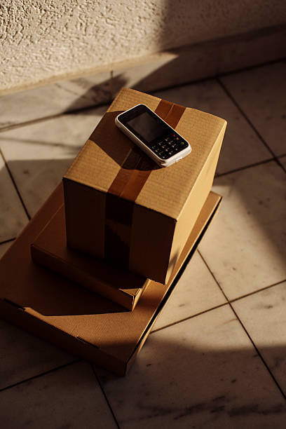 A lone mobile phone sits atop stacked cardboard boxes, bathed in warm sunlight; A simple, everyday scene conveying moving, packing, or delivery moments with a calm, quiet mood