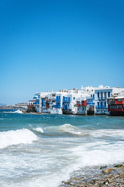 The iconic white and colorful buildings of Little Venice in Mykonos stand right at the water's edge, with waves breaking on a rocky beach.