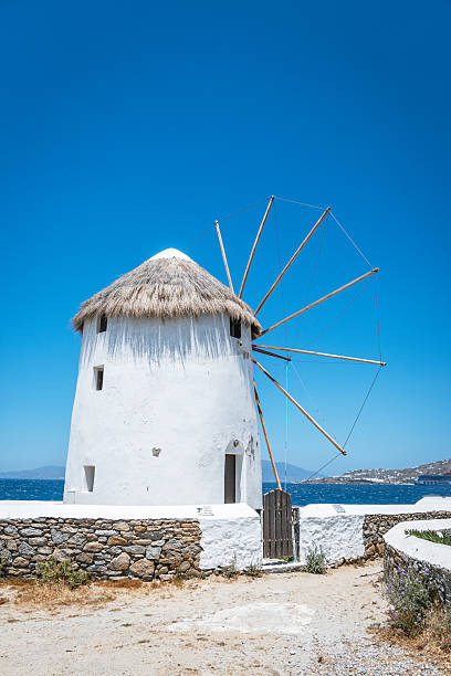 A close-up view of the famous whitewashed Mykonos windmills with their thatched roofs and wooden blades against the backdrop of a clear blue sky and the Aegean Sea.