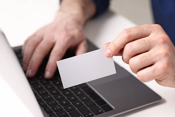 istock Man with laptop holding blank business card at white table, closeup 2244716704
