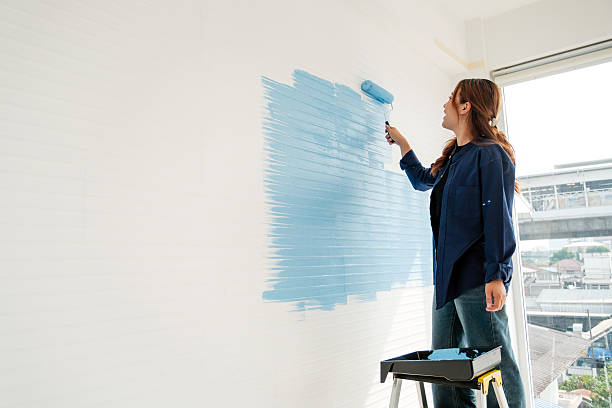 istock Person painting an interior wall light blue with a roller, standing by a step stool and paint tray near a window. 2244727129