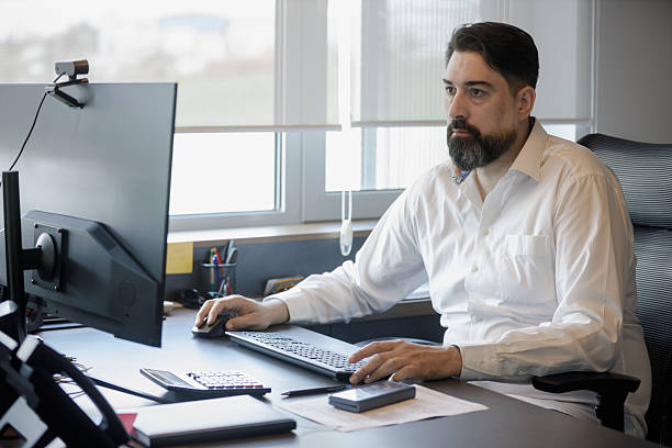 Middle-aged businessman in button down shirt sitting at his office desk and using PC, using mouse and looking at monitor
