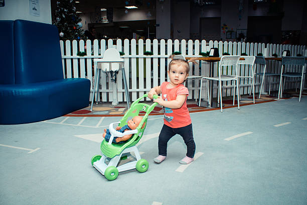 A cheerful toddler girl in a pink t-shirt and pigtails explores a vibrant indoor playroom with ride-on cars, strollers, and interactive toys, expressing curiosity, joy, and growth in a safe and stimulating early childhood environment.