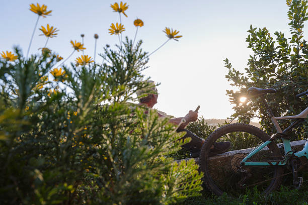 View through wildflowers to mature man sitting on wall, using mobile phone, electric mountain bicycle beside him, Finalborgo