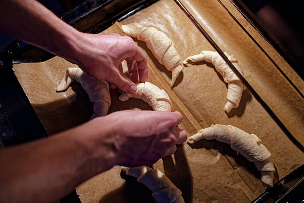 Hands carefully positioning croissant dough on a lined baking tray, illuminated by soft oven light. The image captures the cozy ritual of homemade baking — the scent of flour, the patience before warmth and aroma fill the air and the quiet joy of sustainable, handmade living. Copy space for text, directly above - Food and drink, preparing food