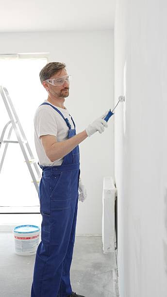 istock Middle aged man wearing protective white gloves and blue work overalls, applying paint with roller on pristine interior wall during home renovation project, vertical view 2244752065