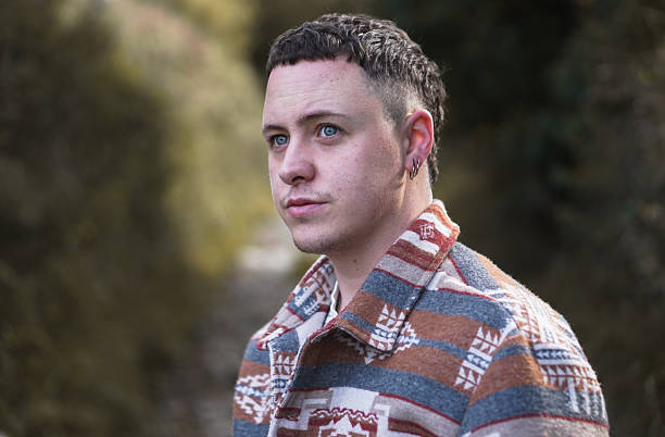 Portrait of a young male with blue eyes standing on a forest path surrounded by autumn tones. The man wears a patterned overshirt in blue, orange, and white colors, gazing thoughtfully into the distance on a cloudy day. Perfect for lifestyle, solitude, nature, mindfulness, and fall season concepts.