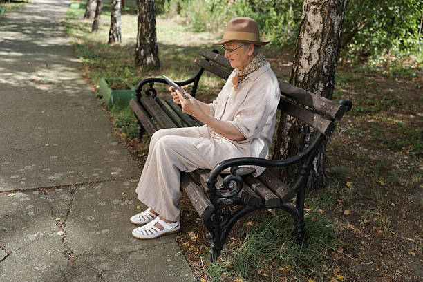 Senior Woman 70+ Sitting On Park Bench Using Digital Tablet Outdoors, Wearing Hat And Scarf, Smiling Calmly While Browsing Online Or Reading. Concept Of Elderly Technology Use, Digital Inclusion, And Modern Lifestyle In Old Age.