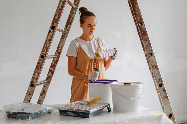 istock A person is preparing to paint a wall using a roller. They stand near a ladder, surrounded by buckets and paint supplies in a bright, airy space 2244760992