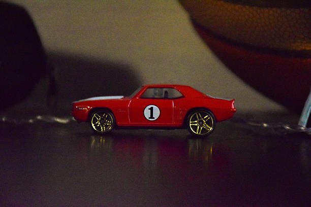 istock Red toy car with golden wheels is positioned on a reflective surface, showcasing its sleek design and vibrant color against a blurred background of sports equipment 2244767640