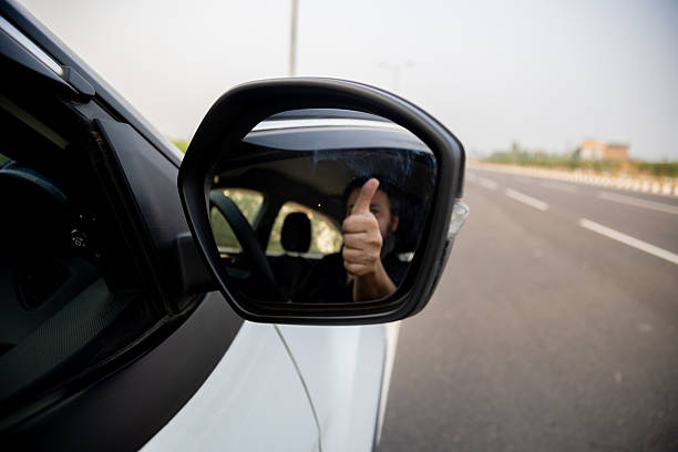 A car driver gives a thumbs-up gesture visible through the rearview mirror, symbolizing confidence, success, and a positive attitude while driving on the road.